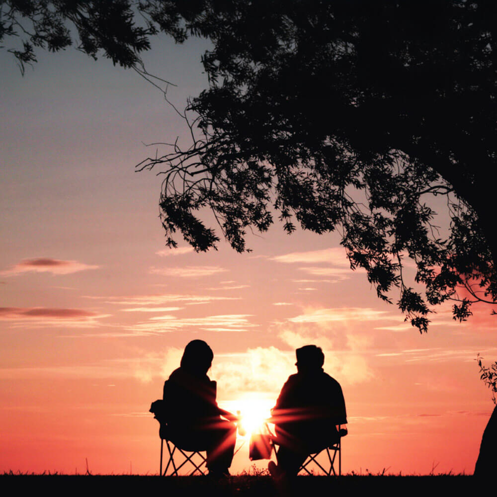 Two people sitting in chairs under a tree at sunset, a couple holding hands on a mountain peak, enjoying the breathtaking view.